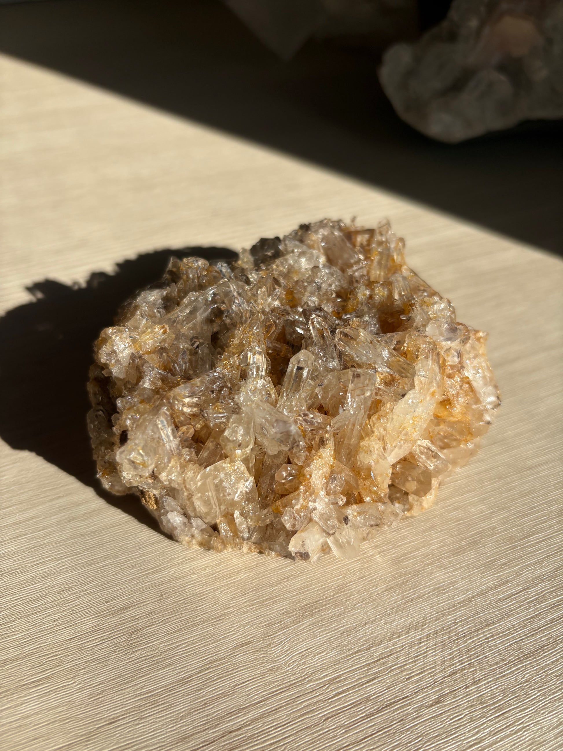 A natural quartz crystal cluster with clear and golden-brown crystalline structures, resting on a light wood surface in direct sunlight.